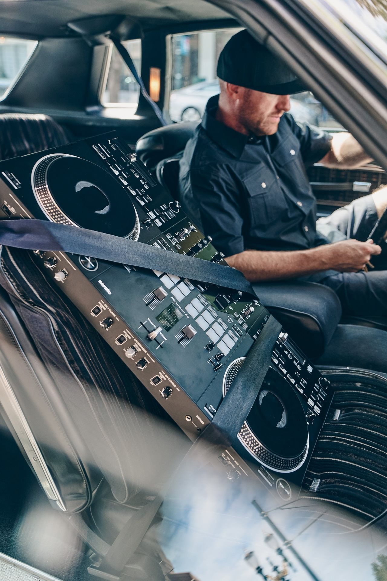 Man in a car with a Pioneer DJ's DDJ-REV7 seat-belted into the seat next to him.